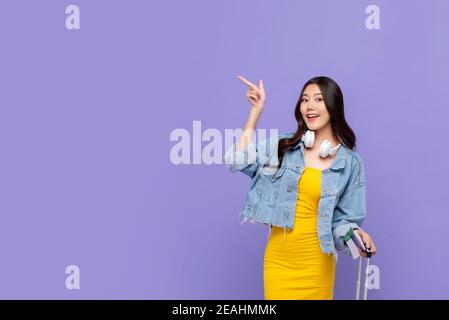 Souriante, jolie femme asiatique touristique prête à partir en voyage en pointant la main pour copier l'espace de côté de l'arrière-plan violet isolé Banque D'Images