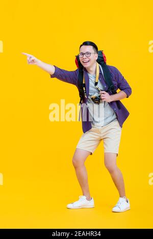 Jeune beau homme asiatique heureux en vêtements touristiques avec sac à dos et des lunettes souriant et pointant tout en tenant l'appareil photo isolé arrière-plan jaune Banque D'Images