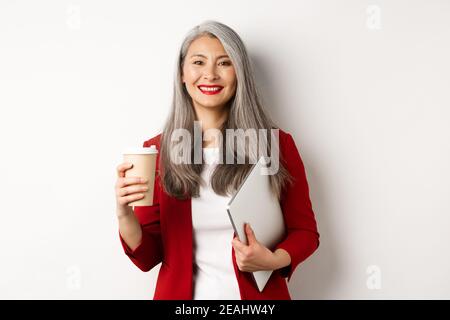 Entreprise. Femme asiatique réussie gérant debout avec une tasse de café et un ordinateur portable, souriant satisfait à l'appareil photo, fond blanc Banque D'Images