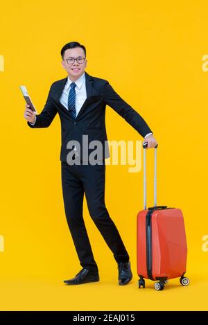 Homme d'affaires asiatique souriant avec une valise tenant un passeport et l'embarquement pass prêt à voyager isolé sur fond jaune Banque D'Images