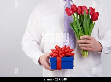 homme adulte dans une chemise blanche et une cravate lilas tenant un bouquet de tulipes rouges avec des feuilles vertes et boîte cadeau Banque D'Images