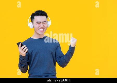 Un homme asiatique émerveillé, portant un casque, écoute ses chansons préférées souriant et accroché son poing isolé sur fond jaune studio Banque D'Images