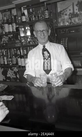 Années 1940, historique, dollars sur le comptoir, comme un homme âgé barman montre deux verres pleins de glace, Oregon, États-Unis. Derrière lui, un bar rempli de bouteilles de spiritueux et une caisse enregistreuse de l'époque, inventée à l'origine en 1879, par James Ritty, propriétaire de saloon à Dayton, Ohio. Banque D'Images