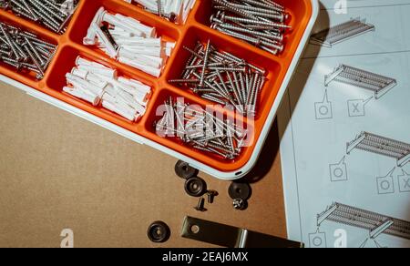 Vis et prise murale dans une boîte en plastique orange. Vue du dessus de la boîte à outils sur fond marron avec instructions de montage du support de séchage de la vaisselle. Jeu de vis et de bouchons en plastique pour le perçage de murs et d'installations. Banque D'Images