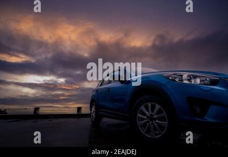 SUV de luxe avec gouttes de pluie. Vue de face nouvelle voiture de SUV bleu garée sur le terrain de stationnement en béton près de la plage de mer avec le ciel de coucher de soleil. Concept de véhicule électrique. Voyage sur route. Voiture couverte de gouttes d'eau. Banque D'Images