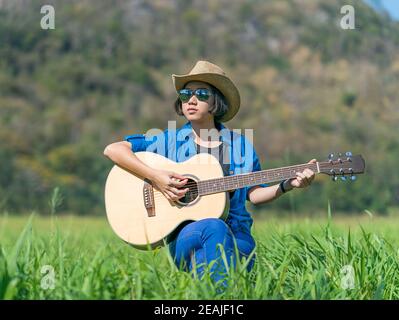 Les femmes cheveux courts porter chapeau et des lunettes de s'asseoir à jouer de la guitare dans grass field Banque D'Images