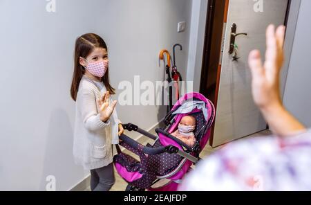 Fille avec masque prêt à aller pour une promenade avec sa poupée avec masque disant au revoir à sa mère Banque D'Images