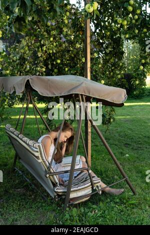 Une adolescente lit un livre assis sur l'oscillation du jardin dans le jardin d'été sous un pommier Banque D'Images