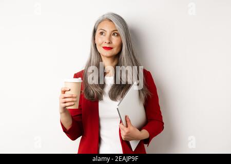 Entreprise. Femme d'affaires asiatique réussie avec cheveux gris, boire du café et debout avec un ordinateur portable, regardant dans le coin supérieur gauche Banque D'Images