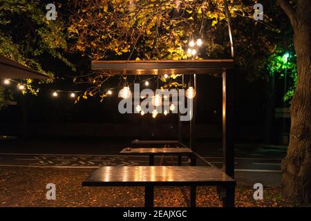 De nombreuses lampes à incandescence différentes sont suspendues sur les tables dans un café de rue dans un parc de nuit Banque D'Images