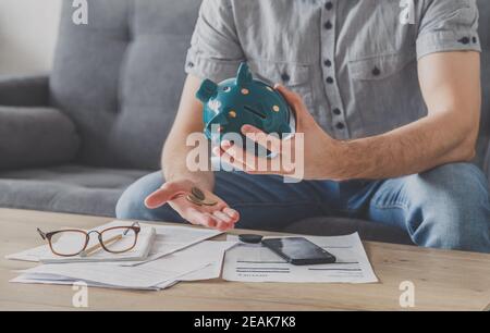 Un homme assis à une table pleine de factures impayées secoue le dernier penny de la banque de piggy. Dépensez vos dernières économies. Chômage, pauvreté, faillite. Banque D'Images