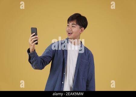 portrait de heureux surpris excité jeune asiatique habillé à la mode je regarde un smartphone isolé sur un fond de studio jaune Banque D'Images