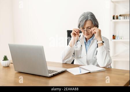 La vieille femme médecin senior avec stéthoscope dans un manteau de laboratoire et des lunettes se fait avec le travail, enlever ses lunettes, assis au bureau et regarder le Banque D'Images