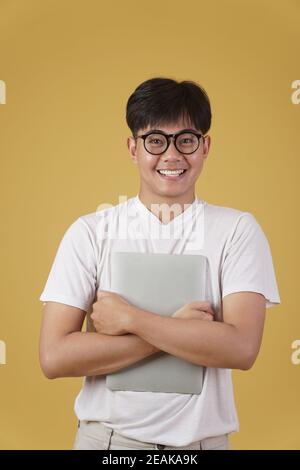 joyeux joyeux jeune asiatique homme nerd étudiant habillé décontracté port lunettes tenant un ordinateur portable isolé sur fond jaune studio Banque D'Images