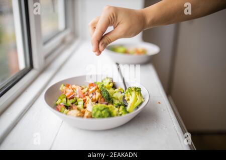 Main ajoutant une pincée de sel à un brocoli et salade de poulet à côté d'une fenêtre Banque D'Images