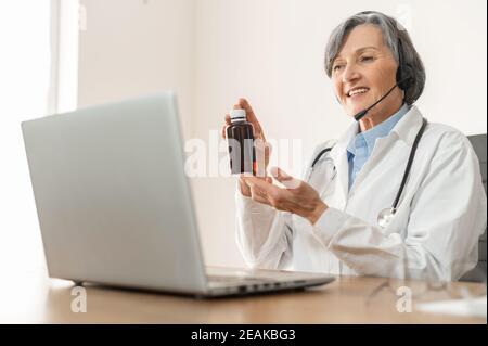 Médecin ou pharmacien de sexe ancien, avec micro-casque dans un blouse de laboratoire, assis devant un ordinateur portable, démontrant la présence d'une bouteille de médicament prescrit au patient Banque D'Images