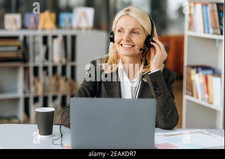 Une femme satisfaite dans son micro-casque et son élégant costume habillé se trouve au bureau et souriant. Élégante femme blonde senior d'affaires travaille dans le service de soutien, de consultation des clients, de communication Banque D'Images