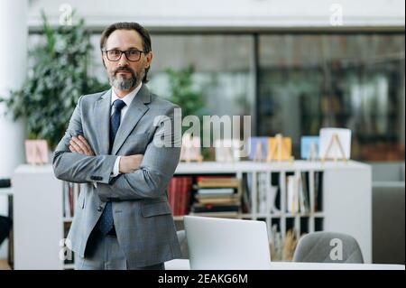 Portrait d'un homme d'affaires caucasien mature et réussi en costume et lunettes formels. Un homme d'affaires influent et confiant se tient debout avec les bras croisés dans un bureau moderne, regarde directement la caméra Banque D'Images