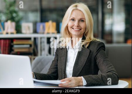 Portrait d'une femme d'affaires heureuse et réussie en tenue officielle sur le lieu de travail. Une femme d'âge mûr et élégante s'assoit à la table au bureau, regarde la caméra avec un sourire amical Banque D'Images