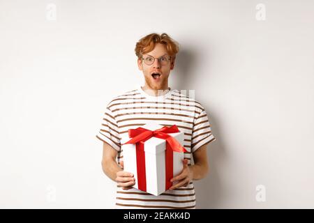 Concept Saint-Valentin et vacances. Surprise redhead petit ami dans les verres regardant reconnaissant à l'appareil photo, recevoir grand présent dans la boîte, debout plus Banque D'Images