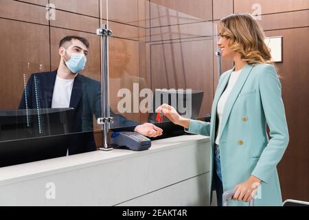 Femme joyeuse tenant la clé près de la réceptionniste dans un masque médical hôtel Banque D'Images