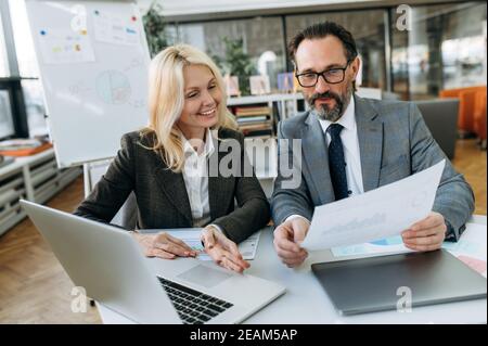 Collègues d'âge moyen et amicaux travaillant ensemble sur le projet, assis à la réception au bureau. Collègues qui réussissent à faire de la paperasserie, à discuter des idées pour le projet, à examiner les graphiques financiers Banque D'Images