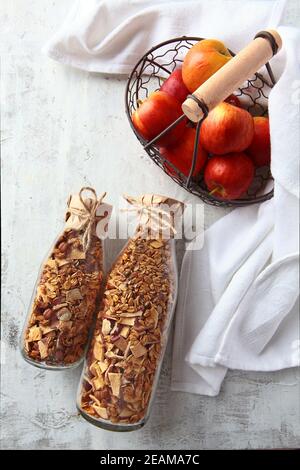 Granola en bouteilles de verre sur fond clair. Pommes rouges dans un panier en métal. Muesli en bouteilles de verre. Une alimentation saine à faible teneur en calories. Produit naturel. Plats végétariens. Photo verticale. Banque D'Images