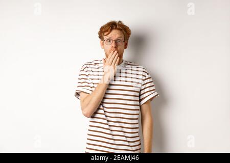 Un jeune homme aux cheveux rouges et aux lunettes entend des nouvelles choquantes ou des persifles, couvre-bouche et regardant l'appareil photo sur fond blanc émerveillé Banque D'Images