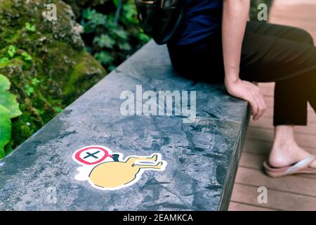 Accent sélectif sur le symbole de l'écart pour la distance sociale afin de prévenir le coronavirus. Concept de distanciation sociale. Femme floue s'asseoir sur le siège près du panneau d'avertissement pour garder la distance sur le siège public au parc extérieur. Banque D'Images