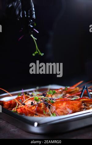 Le chef ajoute des micro-légumes aux crevettes fraîchement cuites. Crevettes géantes d'eau douce grillées. Personne méconnaissable. Mains dans des gants de protection. Concept de cuisine. Banque D'Images