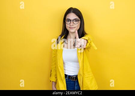Jeune femme d'affaires brune portant un blazer jaune sur fond jaune et de pointer vers le haut avec les doigts numéro un pendant est grave Banque D'Images