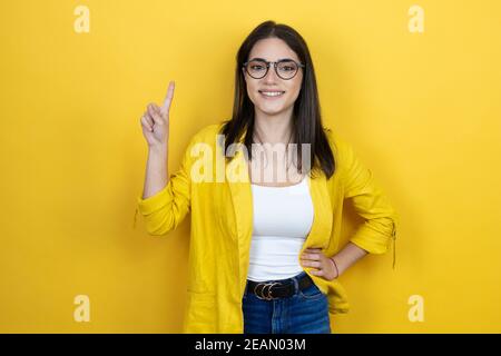 Jeune femme d'affaires brune portant un blazer jaune sur fond jaune et en pointant vers le haut avec les doigts numéro un tout en souriant confiant et Banque D'Images