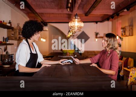 Femme souriante payant avec un smartphone grâce à la technologie NFC. Serveuse tenant un terminal de cartes derrière un comptoir. Payer d'une simple pression dans un café Banque D'Images