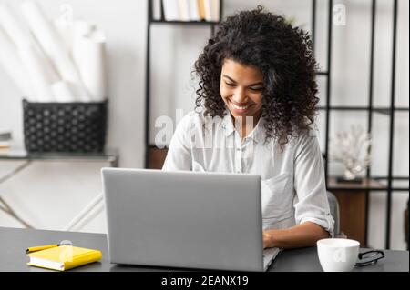 Portrait d'une jeune femme afro-américaine assise à un bureau avec un ordinateur portable, en tapant un e-mail, en envoyant un message à un ami, en discutant, datant en ligne avec un Banque D'Images