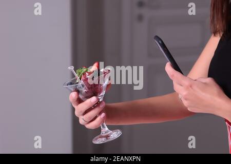 La fille tient dans sa main un verre rempli de différentes baies et prend des photos sur le téléphone. Personne méconnaissable. Buffet à la fête. Dessert dans un verre. Banque D'Images