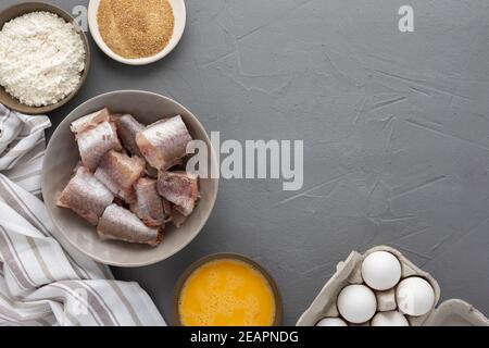 Dans un bol, des morceaux de merlu et des produits pour la cuisine frite, des œufs, de la farine et de la chapelure ont été présentés avec une serviette de cuisine sur fond gris Banque D'Images