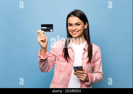 Une jeune femme souriante tient le smartphone et la carte de crédit entre les mains. Photo d'une jeune fille caucasienne portant une tenue à la mode, debout sur un fond bleu isolé, regarde et sourit dans l'appareil photo Banque D'Images