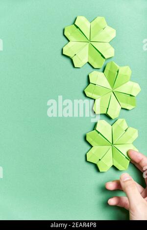 Shamrocks à quatre feuilles d'origami, en papier vert clair, sur fond vert. Une jeune main place un des trèfle au fond. Concepts de luc Banque D'Images