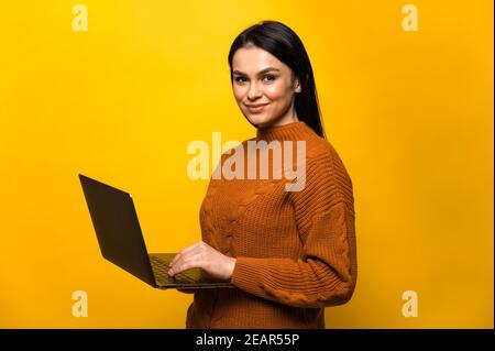 Charmante jeune femme caucasienne souriante dans un chandail décontracté, se tient sur un arrière-plan orange isolé, tient et utilise, regarde et sourit à la caméra Banque D'Images