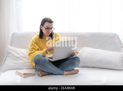 Une fille brune élégante dans un chandail jaune s'assoit à la maison sur le canapé dans une pièce lumineuse et travaille à distance sur un ordinateur portable. Banque D'Images