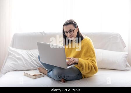 Une fille brune élégante dans un chandail jaune est assise à la maison sur le canapé dans une pièce lumineuse et travaille à distance. Banque D'Images