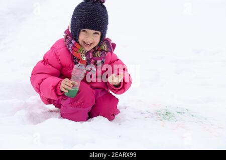 Une jolie fille avec une bouteille de peinture dessine sur la neige. Le concept de la créativité des enfants et du plaisir d'hiver. Banque D'Images