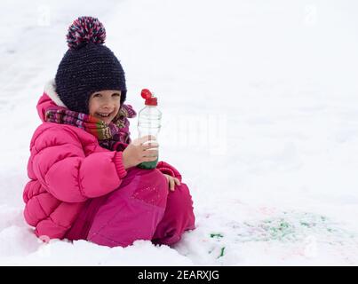 Une jolie fille avec une bouteille de peinture dessine sur la neige. Le concept de la créativité des enfants et du plaisir d'hiver. Banque D'Images