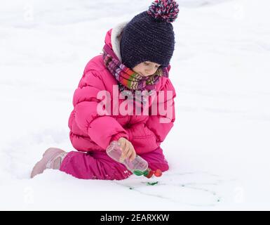 Une jolie fille avec une bouteille de peinture dessine sur la neige. Le concept de la créativité des enfants et du plaisir d'hiver. Banque D'Images