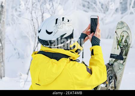 Femme snowboardeuse en costume brillant dans un casque de sport photographié sur un smartphone. Blogueur de voyage. Sports extrêmes. Repos actif. Vue arrière Banque D'Images