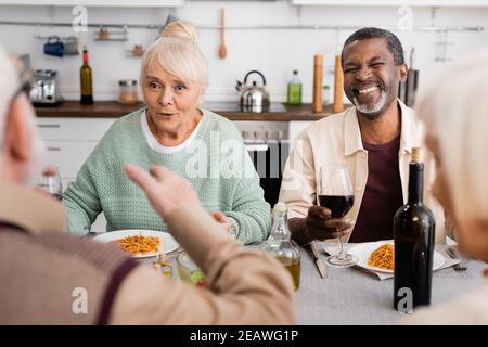 femme âgée stupéfaite regardant l'homme gestant près d'amis interraciaux tout en déjeunant ensemble Banque D'Images