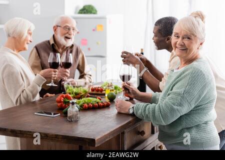 bonne femme âgée tenant un verre de vin rouge et souriant des amis retraités multiculturels sur fond flou Banque D'Images