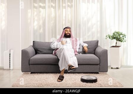 Homme arabe saoudien à la maison se reposant sur un canapé et un aspirateur robot qui nettoie le sol Banque D'Images