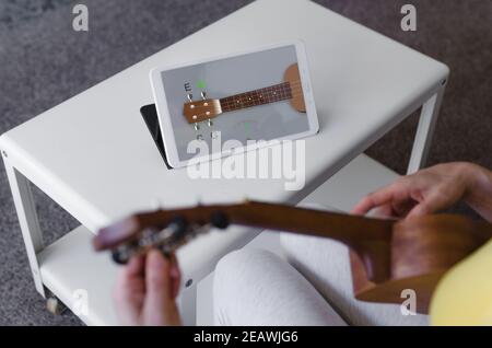 une jeune fille méconnaissable l'ajustant à l'aide d'une application sur sa tablette. Tablette reposant sur une table. Le premier plan avec l'ukulele est hors foyer Banque D'Images