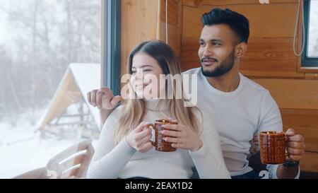 Heureux couple multiracial passant une journée enneigée à Saint-Valentin dans une maison confortable, en buvant du chocolat chaud près de la fenêtre. Photo de haute qualité Banque D'Images